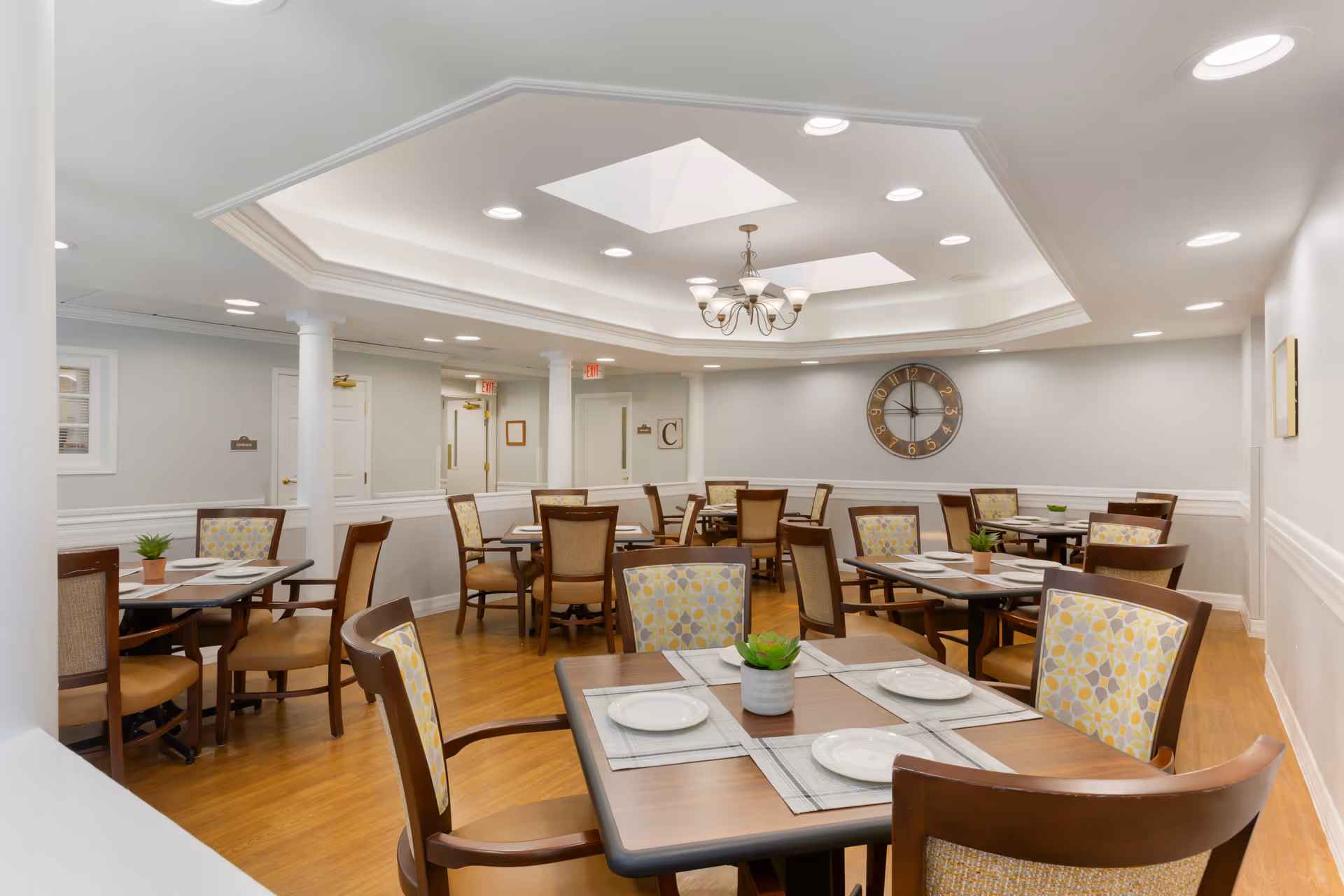 Well-lit dining room with multiple set tables and patterned chairs, a central chandelier and wall clock.