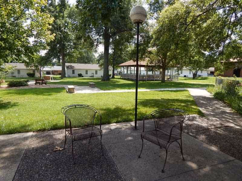 Outdoor seating area with two metal chairs on a concrete patio under shade, surrounded by green grass, trees, and a gazebo in the background at a senior living facility.