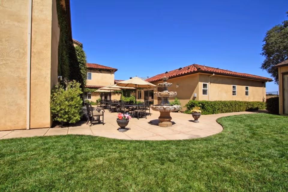 Outdoor patio area at New West Haven facility with a multi-tiered stone fountain in the center, surrounded by tables and chairs with beige umbrellas. The patio is bordered by green grass and beige stucco buildings with red tile roofs under a clear blue sky.
