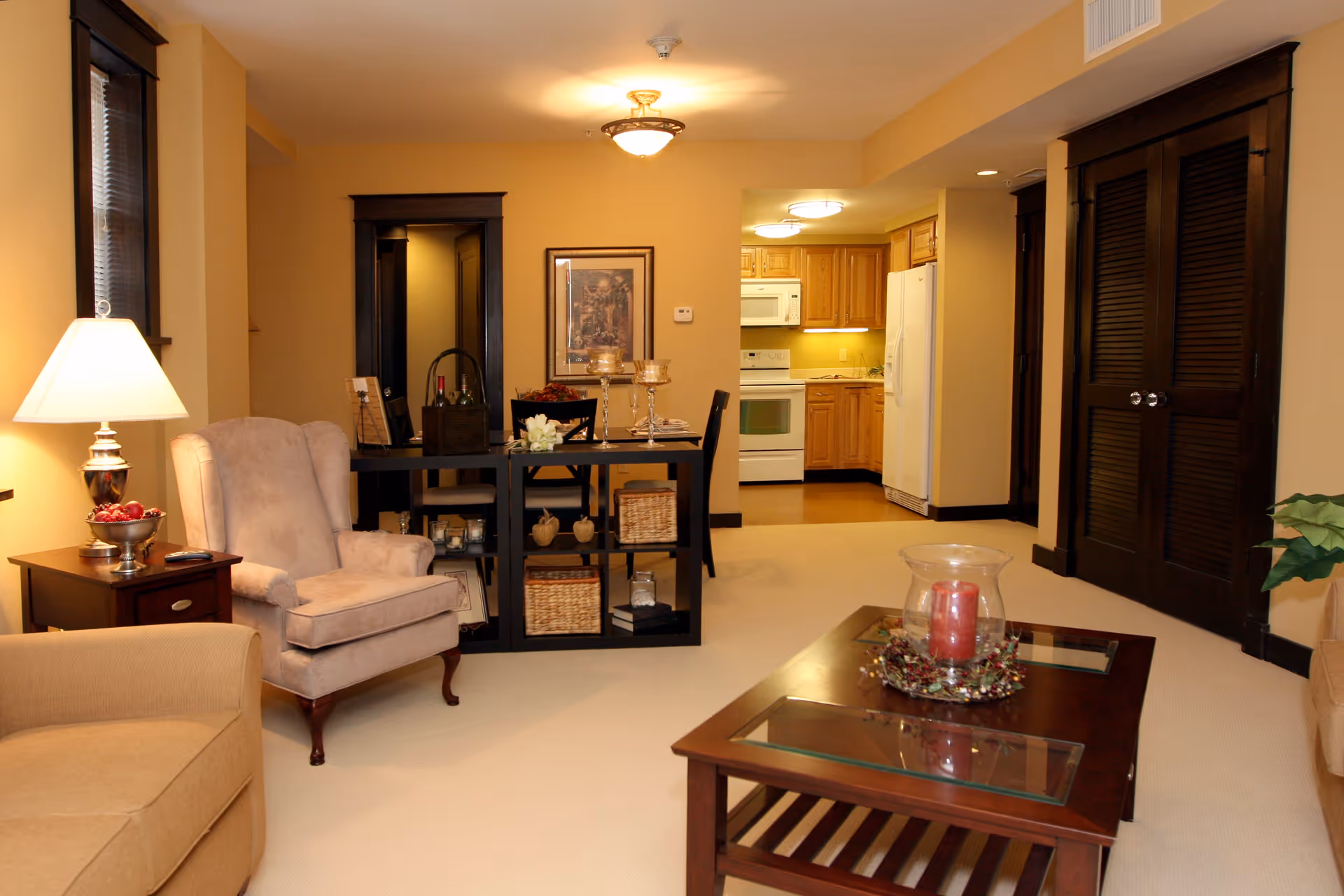 A cozy living room and dining area with beige walls and carpet. The living room features a beige armchair, a beige sofa, a wooden coffee table with a red candle centerpiece, and a side table with a lamp and decorative bowl. The dining area has a black dining table with chairs and decorative items. In the background, there is a kitchen with wooden cabinets, a white refrigerator, stove, and microwave. Dark wood trim and doors contrast with the light-colored walls and carpet.