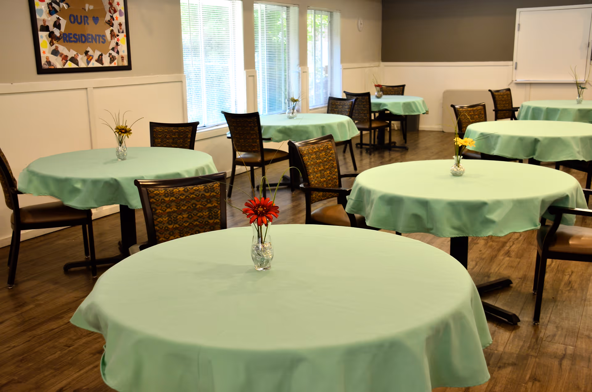 A dining room with several round tables covered with light green tablecloths, each table has a small vase with flowers. The room has wooden flooring, multiple chairs around the tables, large windows letting in natural light, and a bulletin board on the wall with a sign that reads 'Our Residents'.