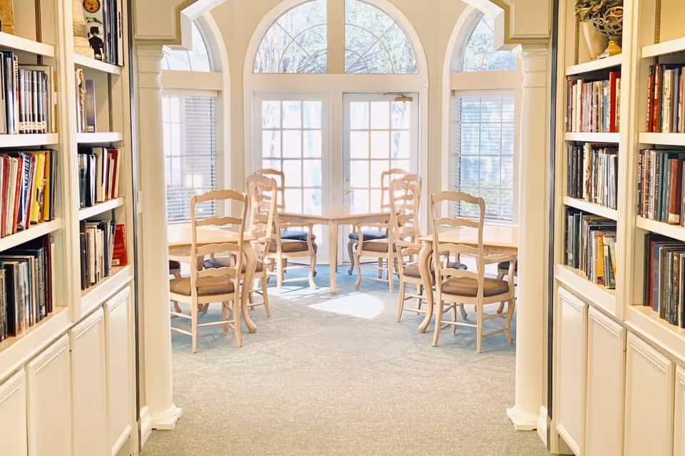 A bright room with large windows and glass doors letting in natural light. The room has several wooden chairs with cushioned seats arranged around small tables. On either side of the entrance to the room, there are built-in bookshelves filled with books. The walls and furniture are light-colored, creating a warm and inviting atmosphere.