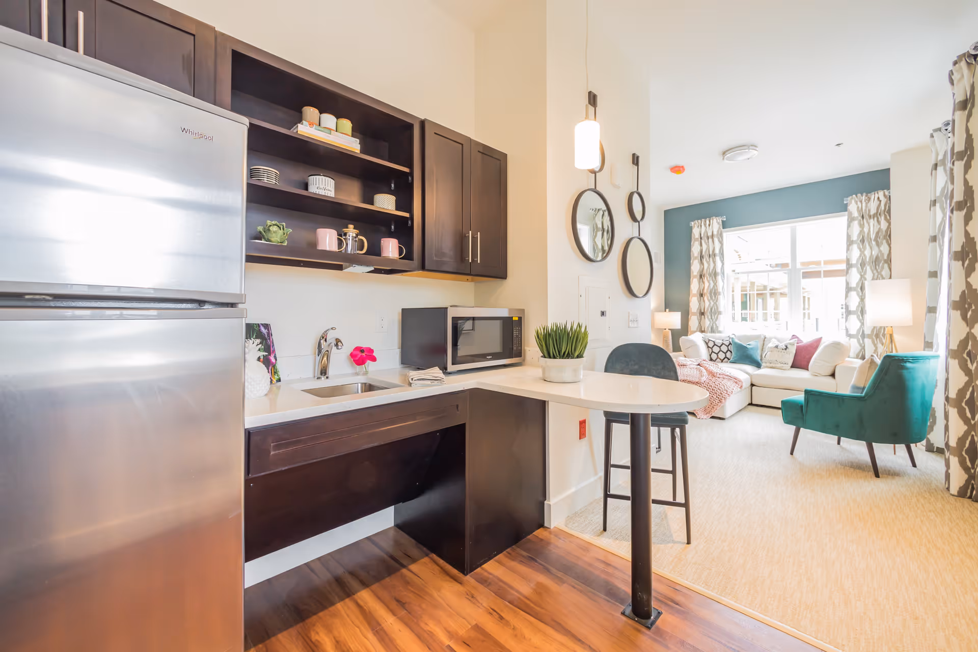 Open kitchenette with a stainless refrigerator, dark cabinets, a small bar counter and stool opening into a bright living room with a sofa and teal armchair.