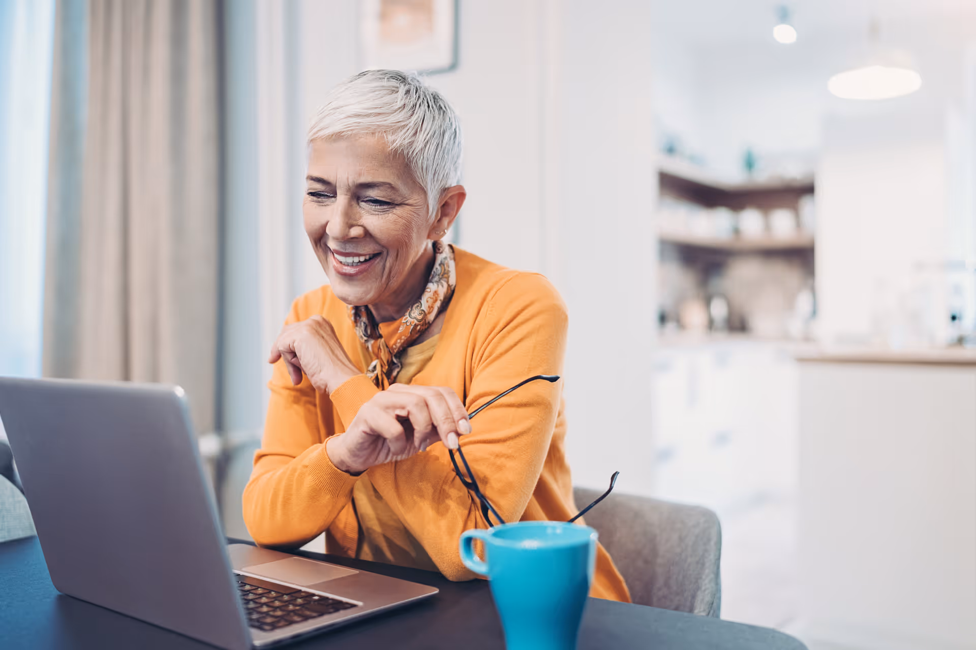 An elderly woman with short white hair wearing an orange sweater and a patterned scarf is sitting at a table, smiling and looking at a laptop. She is holding a pair of glasses in one hand, and there is a blue mug on the table in front of her. The background shows a bright, modern kitchen area.