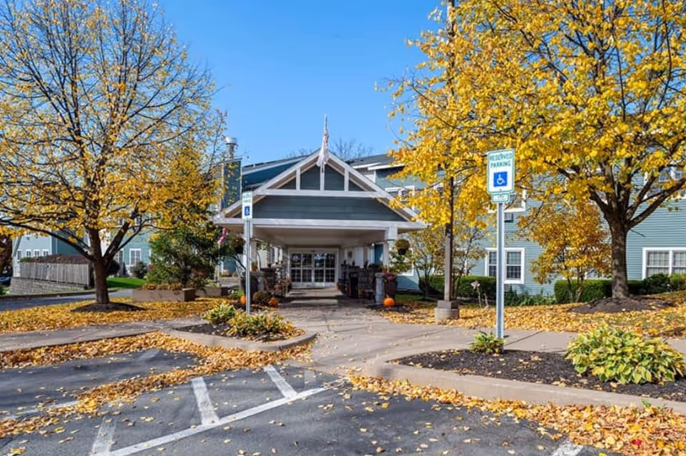 Front entrance of a senior living building with a covered entryway, fall trees with yellow leaves, and accessible parking signs.