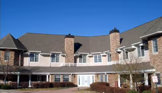 Exterior view of a two-story senior living facility building with beige siding and brick accents, multiple windows, two prominent chimneys, and a covered entrance. The sky is clear and blue, and there are some small trees and bushes in front of the building.
