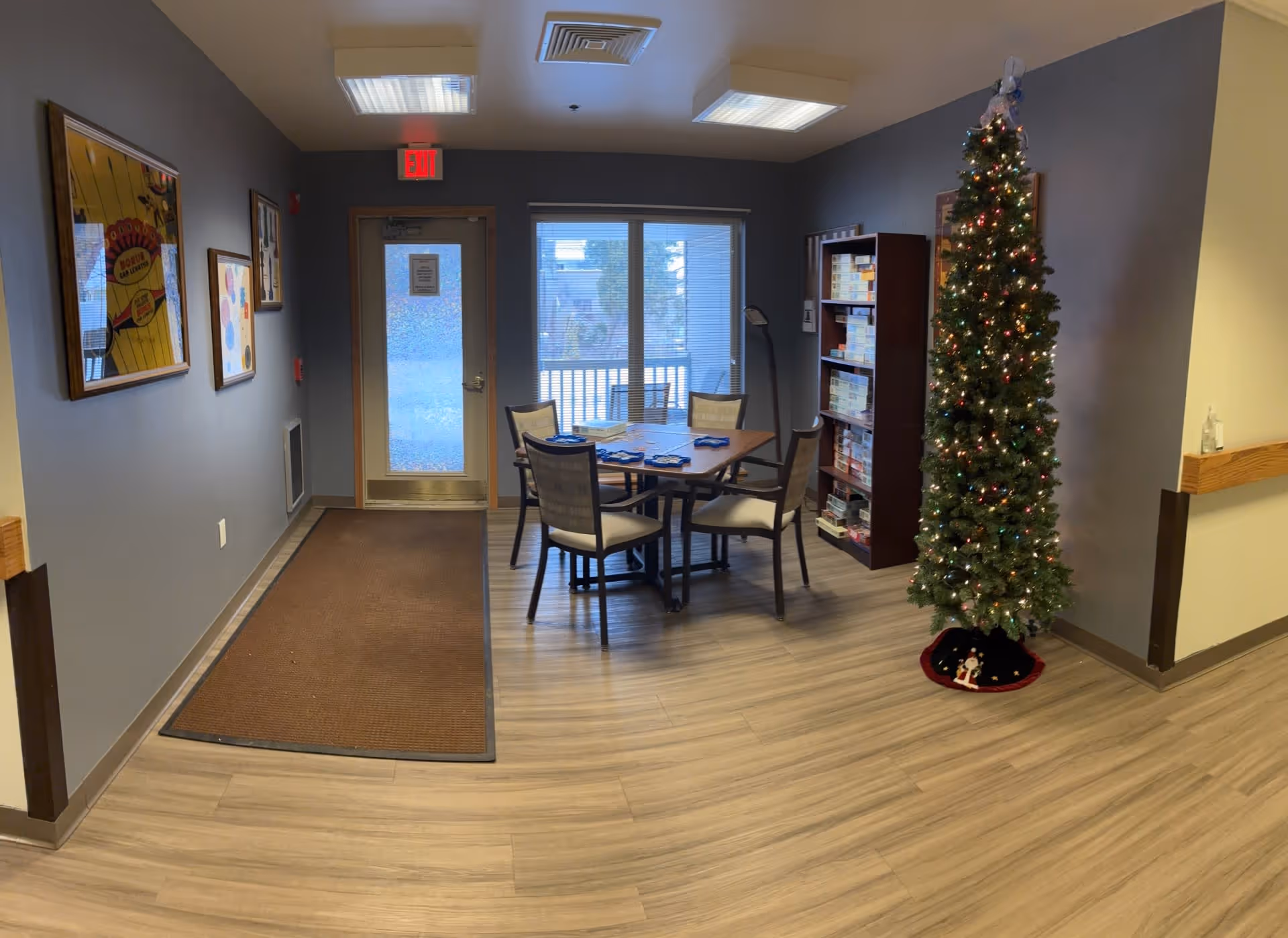Interior view of a common area in a senior living facility with a decorated Christmas tree on the right, a table with four chairs in the center, a bookshelf filled with board games, and a door with a window and an exit sign above it. The walls are painted gray and there are framed pictures hanging on the left wall.