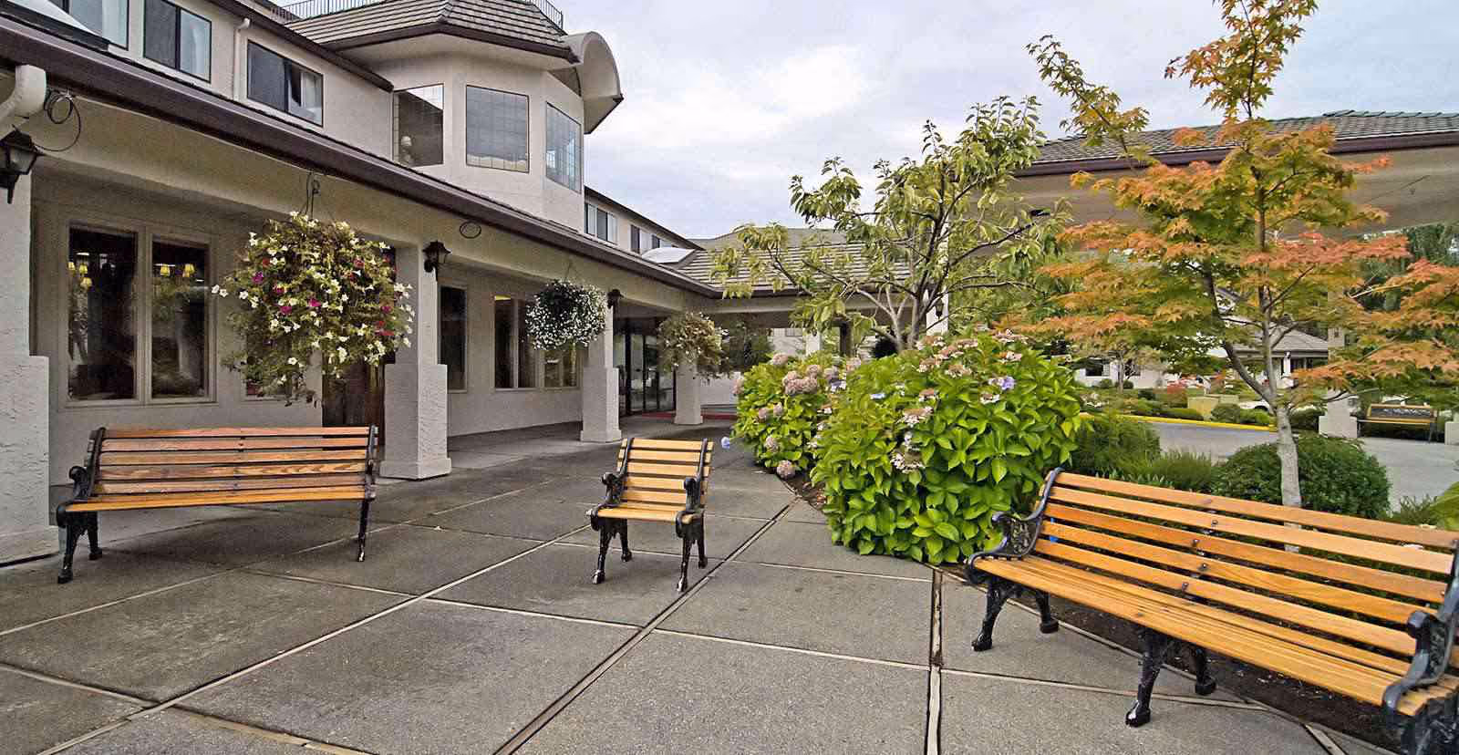 Entrance courtyard of a senior living building with wooden benches, hanging flower baskets, and landscaped shrubs.