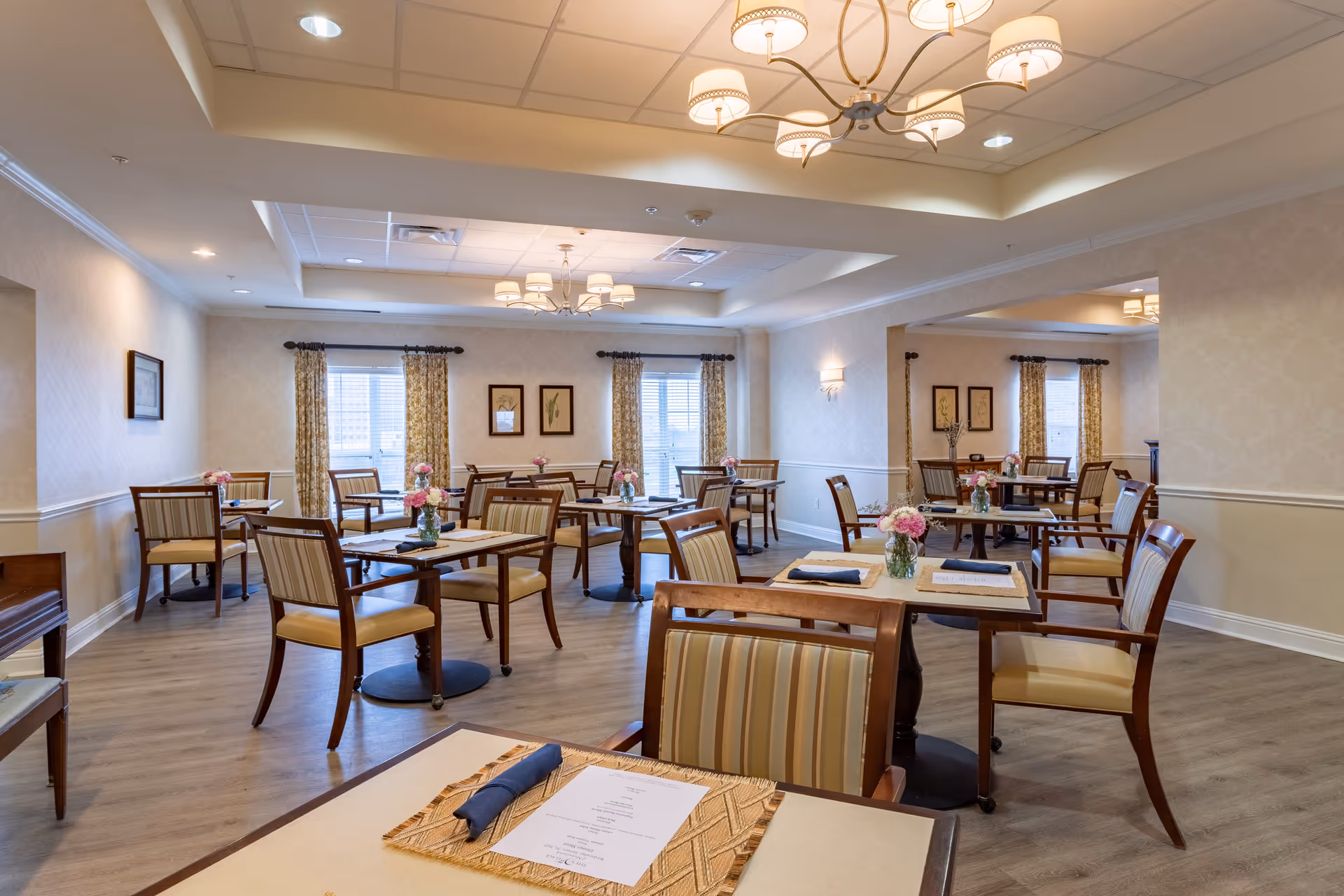 Interior view of the dining room at The Village of Meyerland, featuring wooden tables with striped chairs, floral centerpieces, and large windows with curtains.