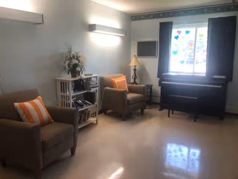 A small living room area with two cushioned armchairs, each with an orange and white striped pillow. There is a small bookshelf with books and a flower arrangement on top, a standing lamp, and a window with dark curtains letting in natural light. A piano is positioned below the window.