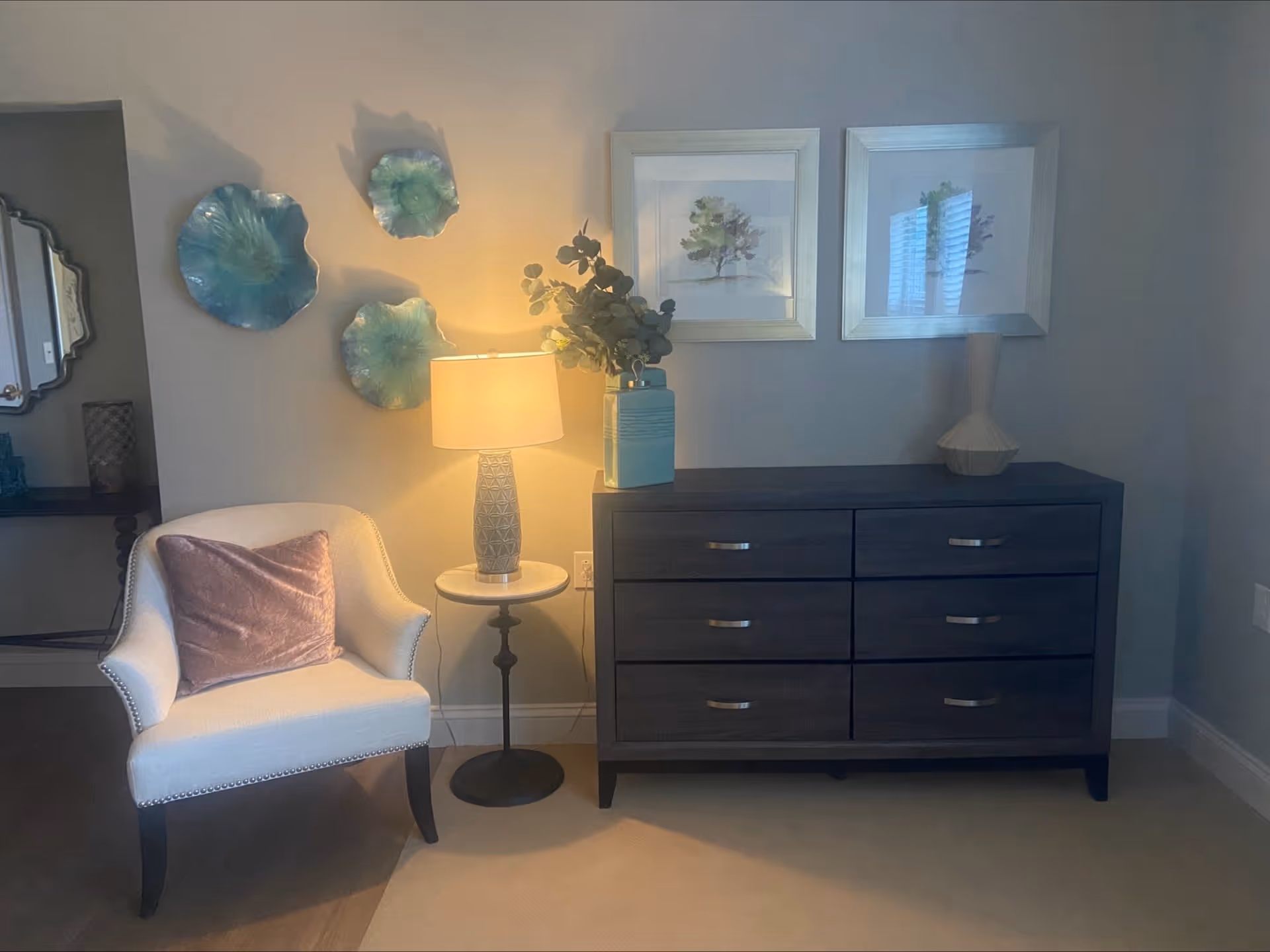 A cozy corner of a room featuring a white upholstered armchair with a pink velvet pillow, a small round side table with a decorative lamp, and a dark wooden dresser with six drawers. Above the dresser are two framed botanical prints and a ceramic vase with greenery. Three decorative blue-green wall hangings are displayed on the wall beside the lamp.