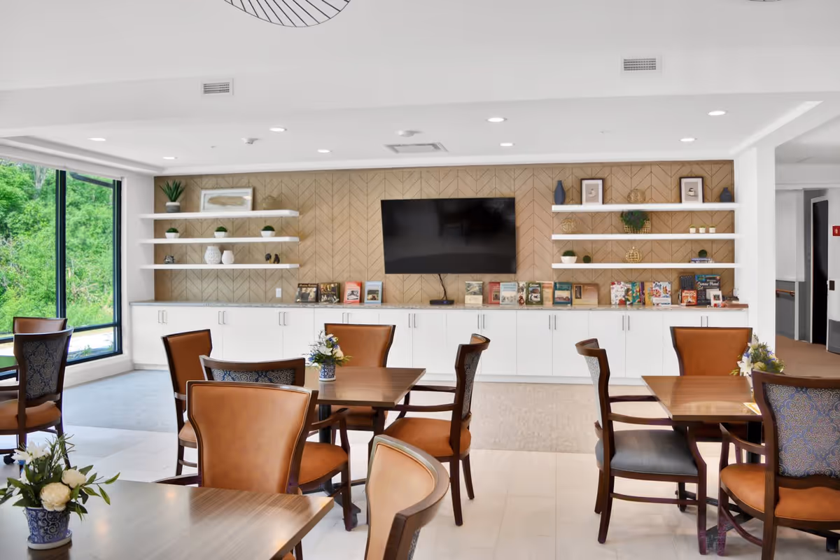 A bright and modern common area with several wooden tables and chairs arranged neatly. The back wall features white cabinets with a marble countertop, open white shelves decorated with plants, vases, and books, and a large flat-screen TV mounted in the center. Large windows on the left side allow natural light to fill the room and offer a view of green trees outside.