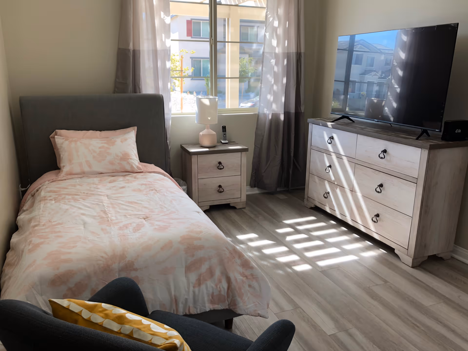 A bright bedroom with a single bed covered in a light pink and white patterned comforter and pillow. Next to the bed is a small wooden nightstand with a lamp and a phone. Across from the bed is a wooden dresser with a large flat-screen TV on top. Sunlight streams through a window with sheer curtains, casting shadows on the floor.