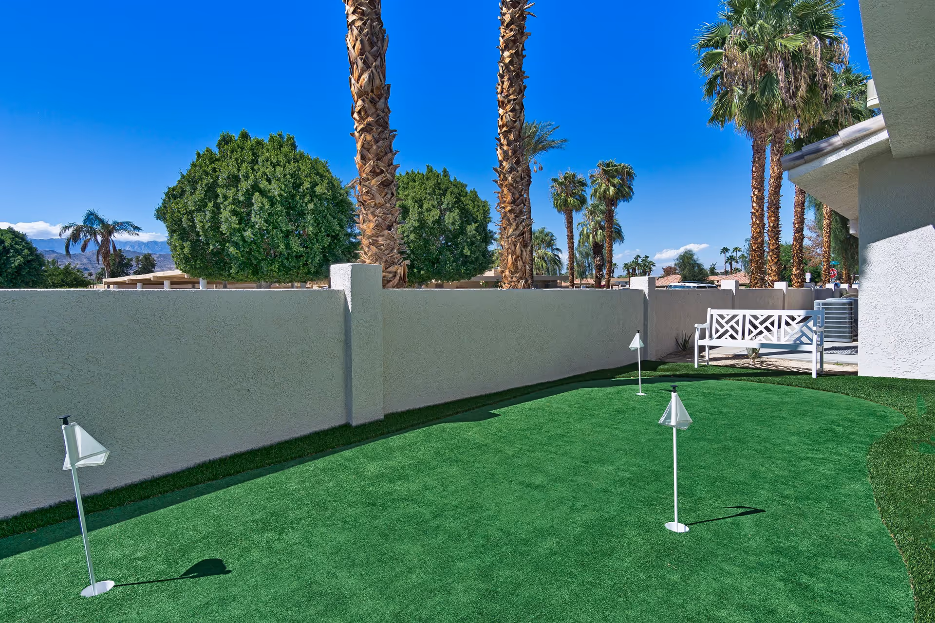 Small outdoor putting green with practice flags, surrounded by a stucco wall, palm trees, and a white bench.