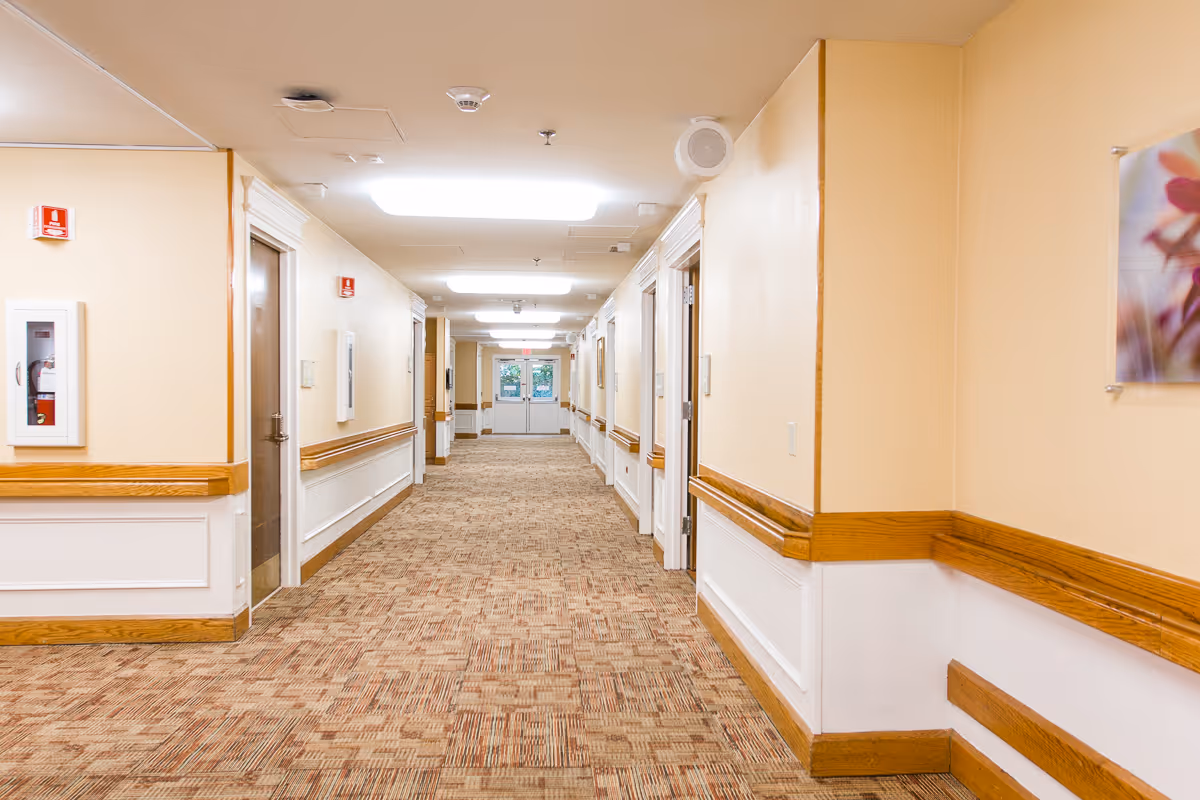 A long, well-lit hallway in a senior living facility with beige walls, wooden handrails, and patterned carpet. Several doors line both sides of the hallway, and double exit doors are visible at the far end. A fire extinguisher is mounted on the left wall.