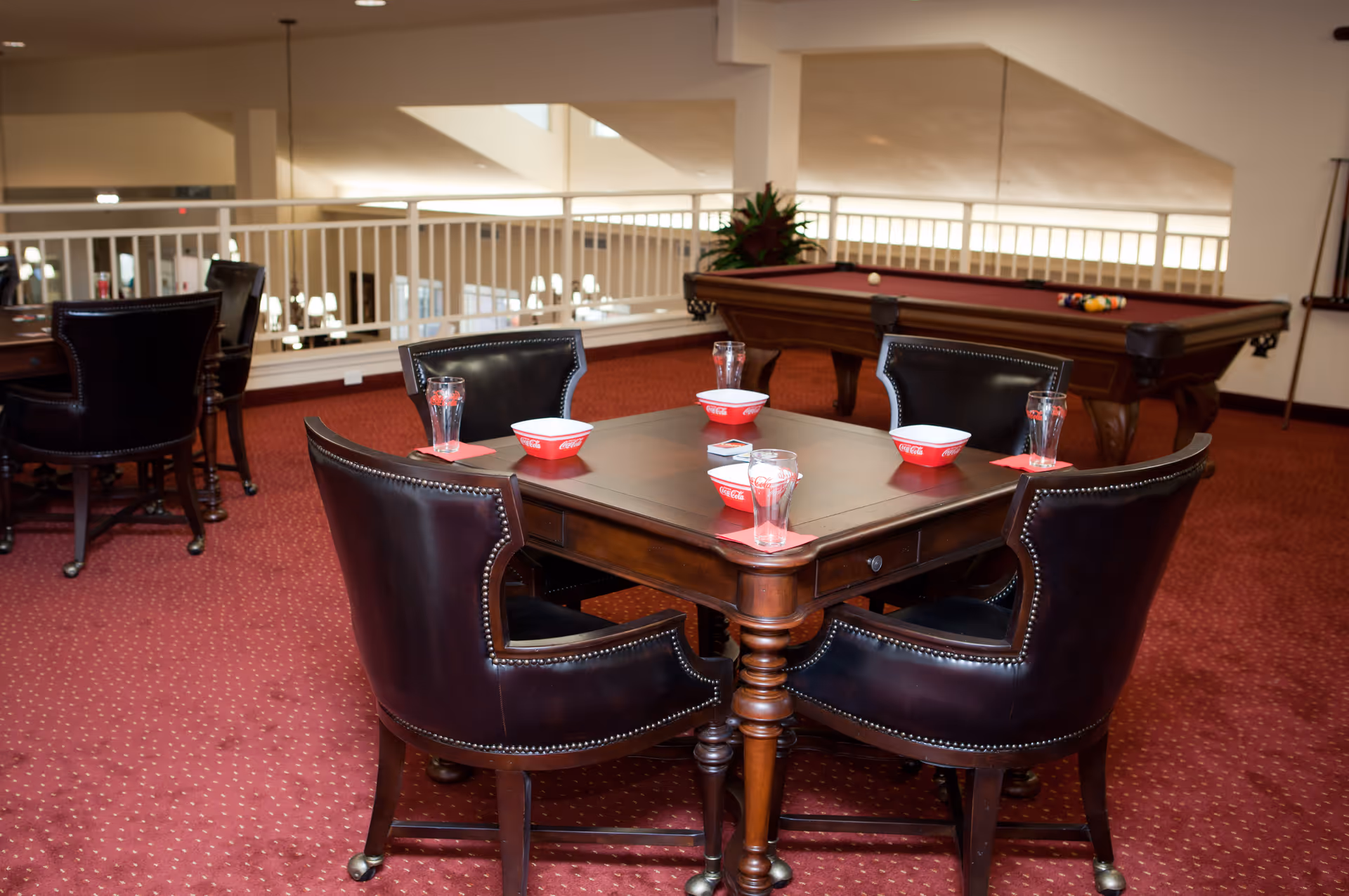 A cozy indoor recreational area with a wooden table surrounded by four black leather chairs. On the table, there are four glasses and four red bowls placed on red napkins. In the background, there is a pool table with billiard balls arranged on it, and the area has red carpeting and white railings.