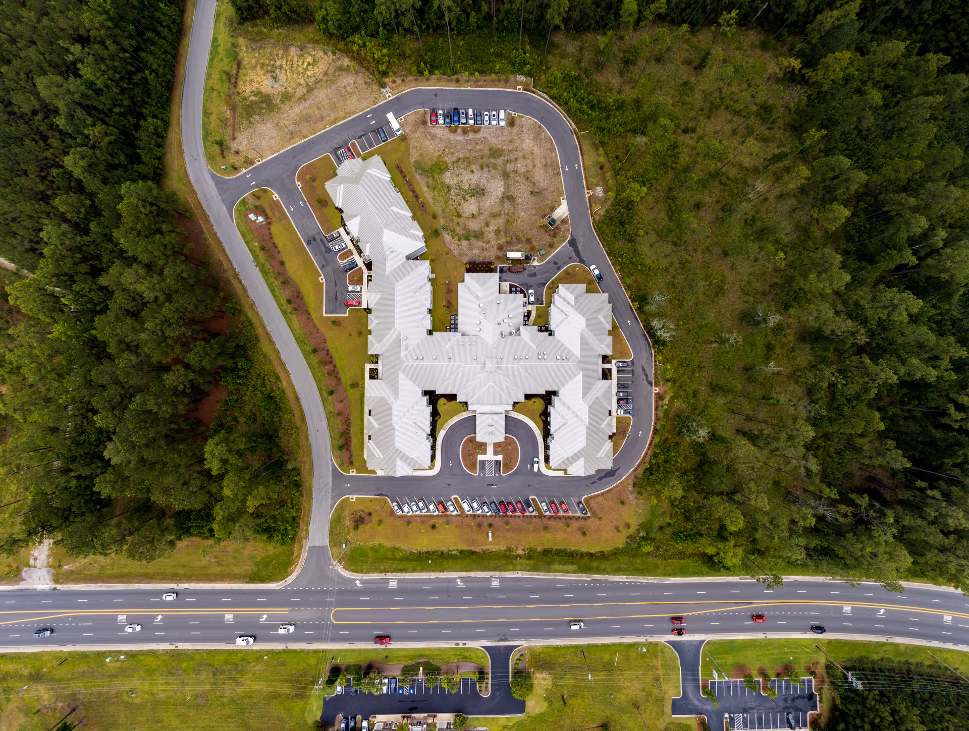Aerial view of a large senior living facility named Arbor Landing at Surfside surrounded by trees and greenery, with a curved driveway and parking areas around the building, adjacent to a multi-lane road with vehicles.