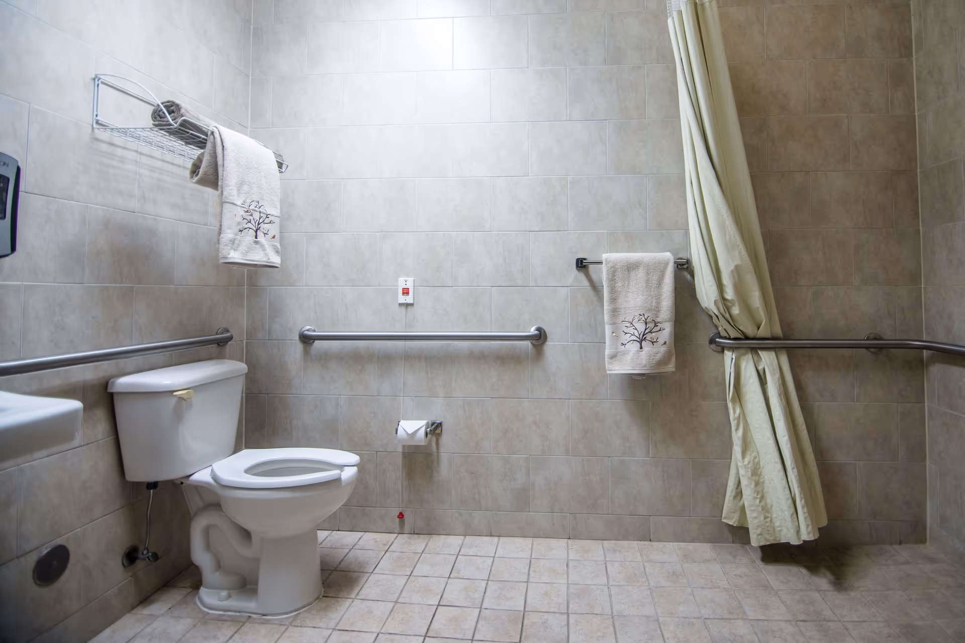 A clean and accessible bathroom with beige tiled walls and floor, featuring a white toilet with grab bars on the walls, a towel rack with a towel, a toilet paper holder, and a shower area with a curtain and grab bars.