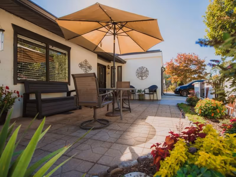 Sunlit patio with a table, chairs and a large umbrella next to a building entrance, bordered by potted plants and colorful flower beds.