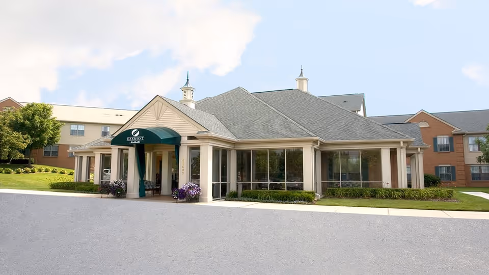 Exterior view of Oakmont Northville senior living facility showing a single-story entrance building with large windows, a green awning over the entrance, and surrounding landscaping with grass and shrubs. The sky is partly cloudy.