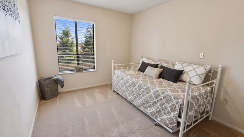 A small bedroom with a single white metal daybed covered with a beige and white patterned quilt and several pillows. There is a window with blinds letting in natural light, a small plant on the windowsill, and a laundry basket with a blanket in the corner. The walls are light-colored and the floor is carpeted.