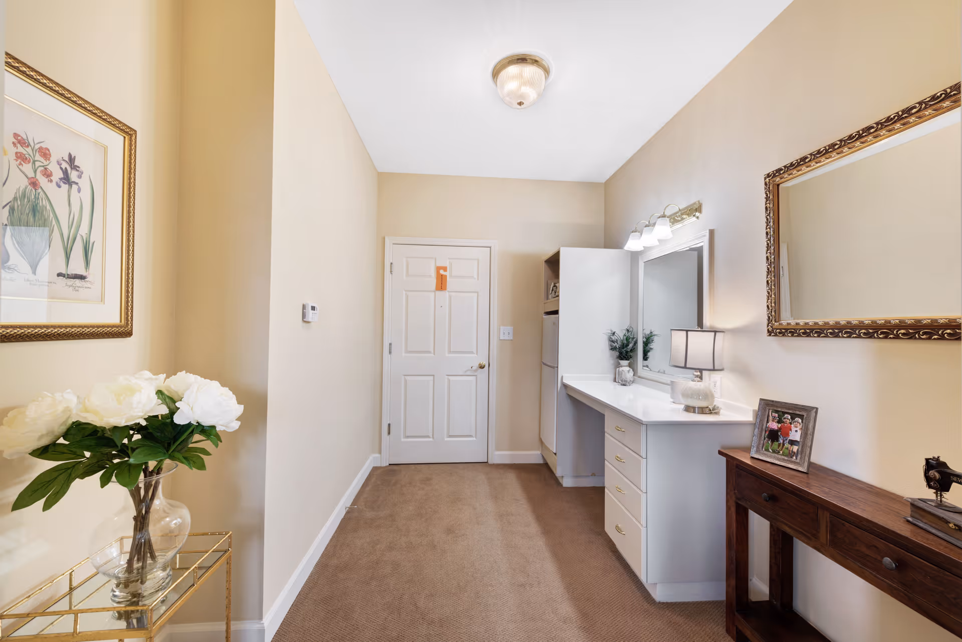 A hallway area in a senior living facility with beige walls and carpeted floor. On the left side, there is a glass table with a vase of white flowers and a framed floral artwork above it. At the end of the hallway is a white door. On the right side, there is a white vanity desk with drawers, a mirror with lights above it, a small lamp, and a decorative plant. Next to the vanity is a wooden console table with a framed photo and a small decorative item. A large framed mirror hangs above the console table.