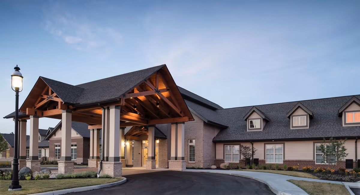 Exterior view of Liberty Place Memory Care facility at dusk, showing a large covered entrance with wooden beams, brick and siding walls, multiple windows, and a curved driveway with landscaping and a street lamp.