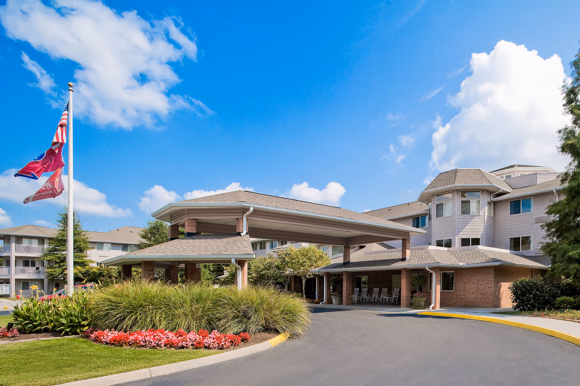 Exterior view of Holiday Echo Ridge senior living facility showing the main entrance with a covered driveway, landscaped garden with flowers and shrubs, flagpole with flags, and a multi-story building under a blue sky with some clouds.