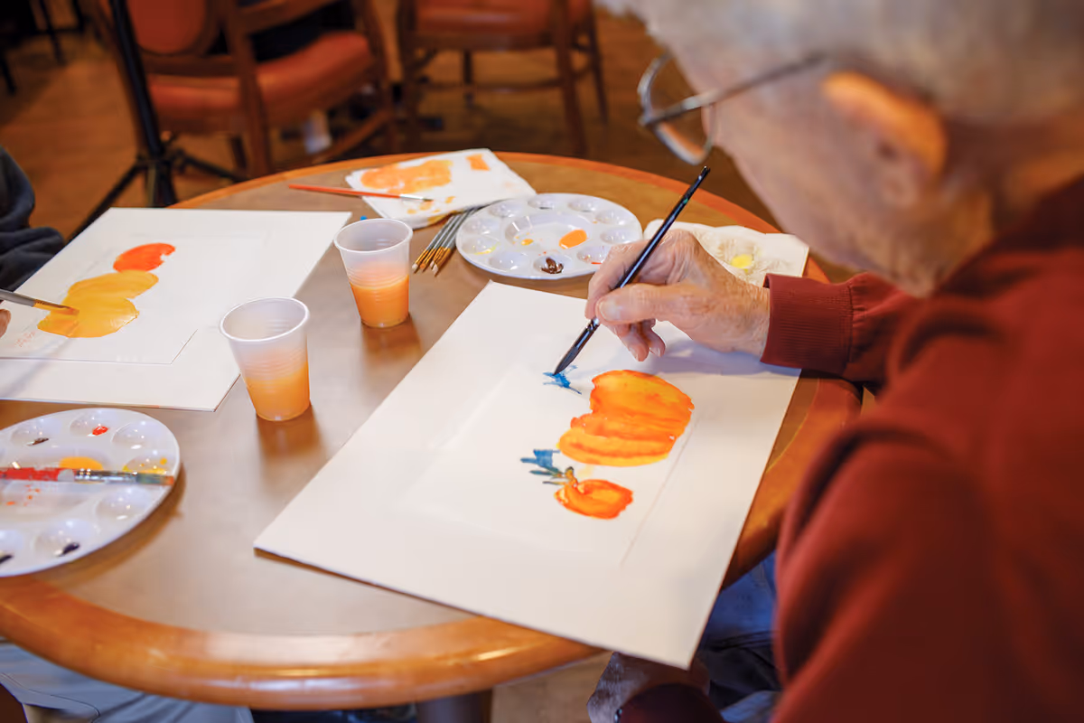 An elderly person painting a picture of orange pumpkins on white paper at a round wooden table. There are paint palettes, brushes, and plastic cups with orange liquid on the table. Another person is partially visible also painting at the same table.