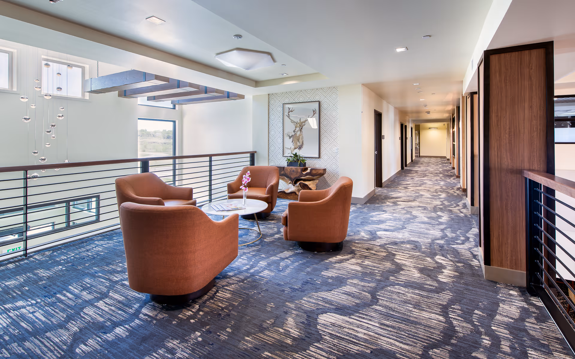 A modern indoor seating area in a senior living facility with four orange armchairs arranged around a small round table with a flower vase. The space features large windows, a decorative hanging light fixture, patterned carpet, and a hallway with multiple doors leading to rooms. A framed artwork of a deer is mounted on the wall.