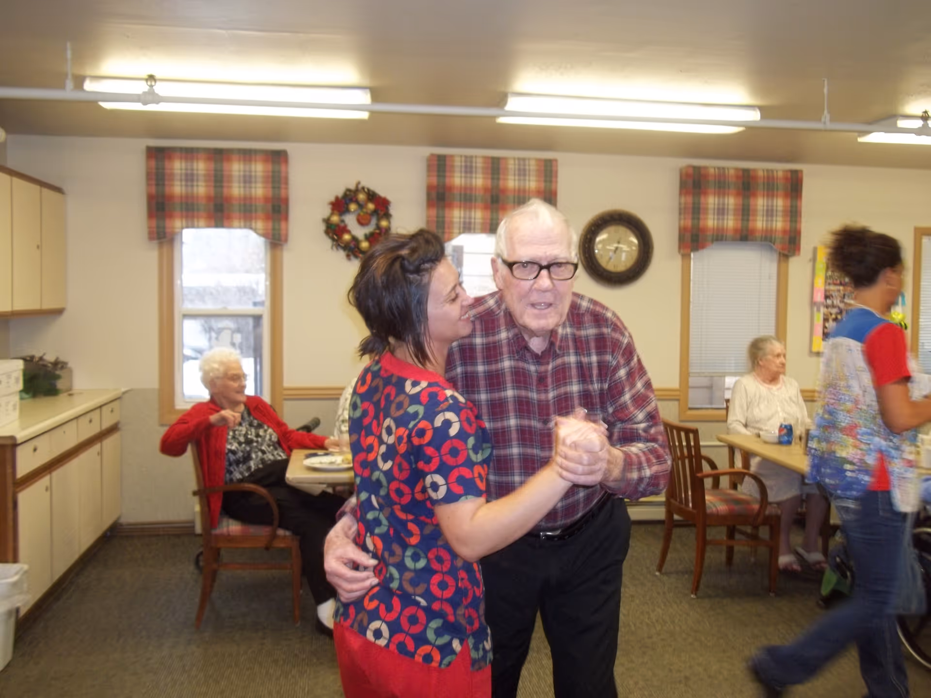 An elderly man and a caregiver dance together in a communal dining room while other residents sit at tables.