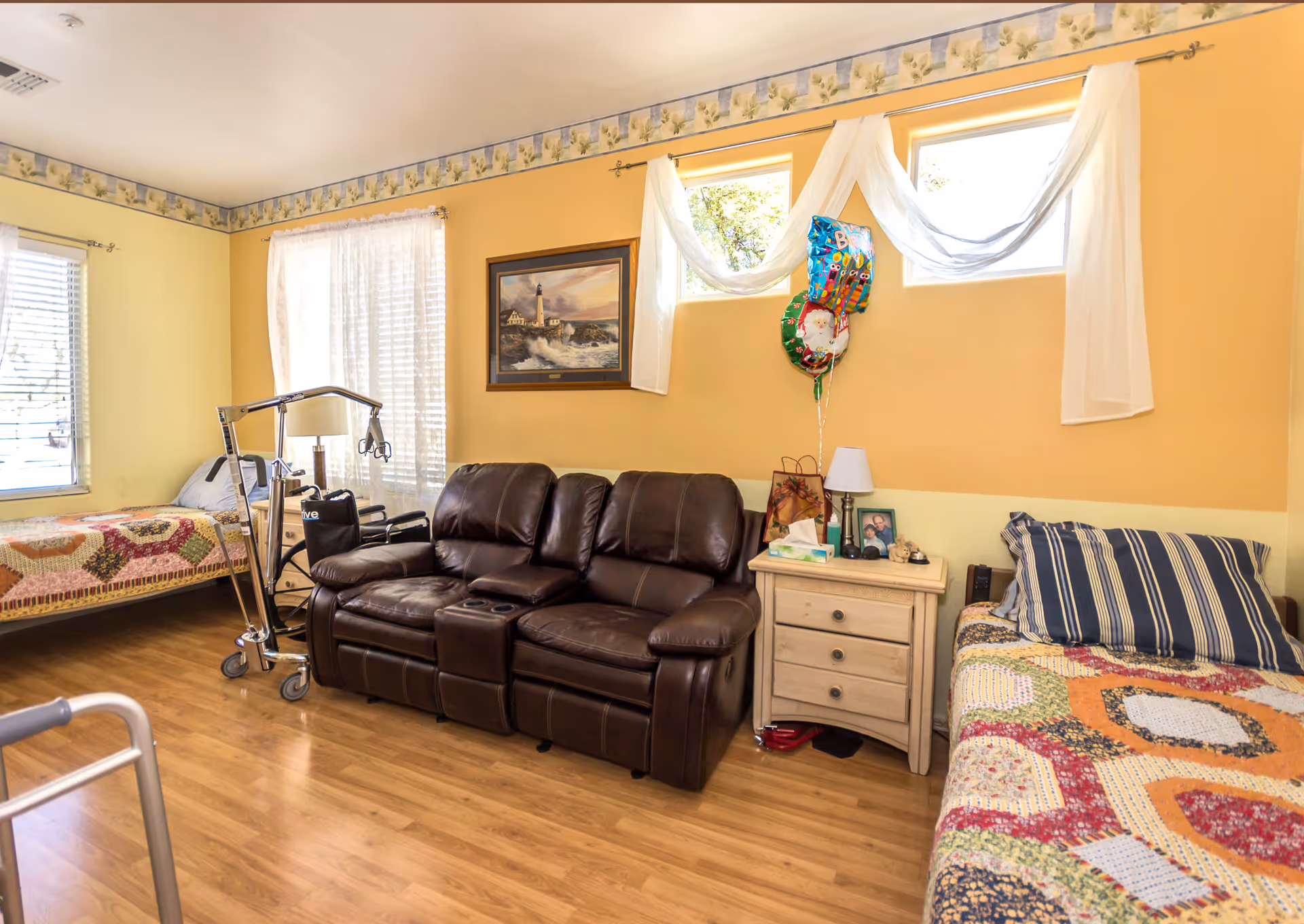 A cozy room with two single beds covered in colorful patchwork quilts, a dark brown leather loveseat in the center, and a wooden nightstand with a lamp, tissue box, and framed photo. The walls are painted yellow with a floral border near the ceiling, and there are three windows with sheer white curtains. A walker and a patient lift are also visible in the room.