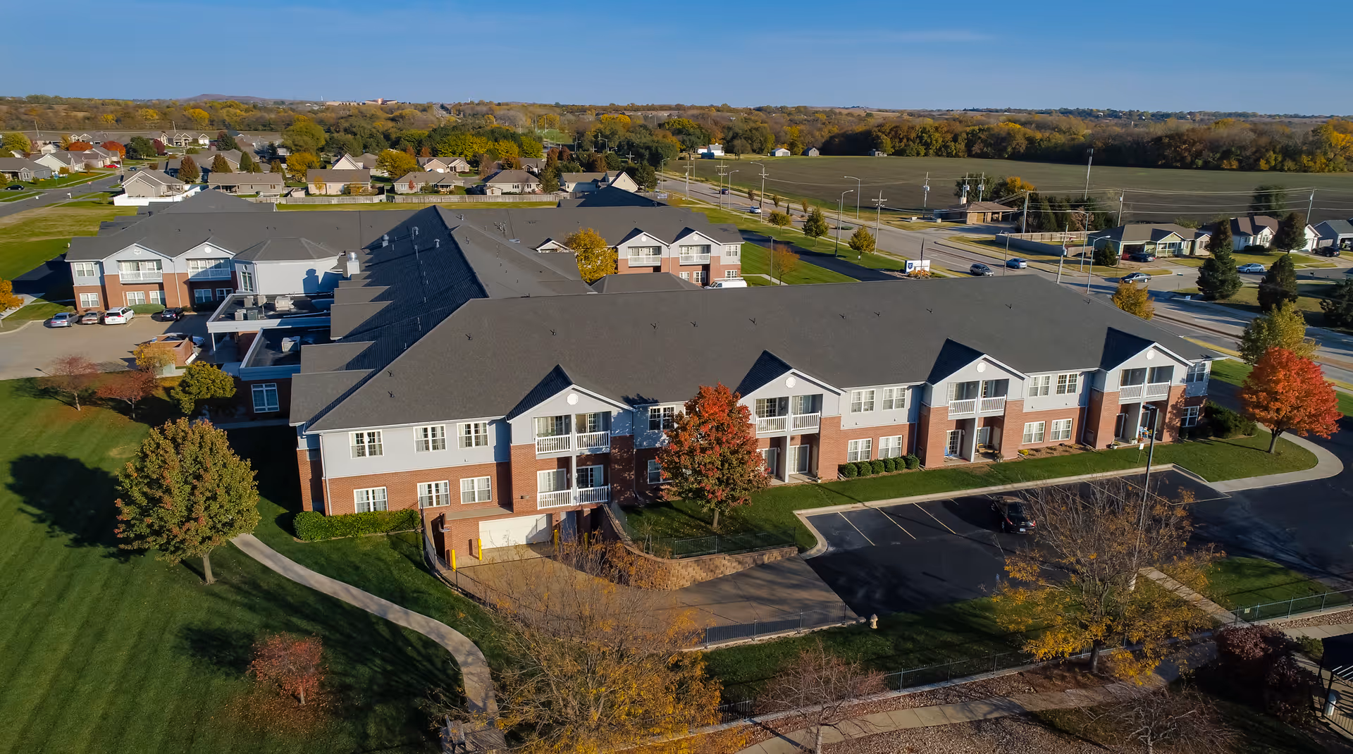Aerial view of a two-story brick-and-white senior living building with parking, lawns, and trees.