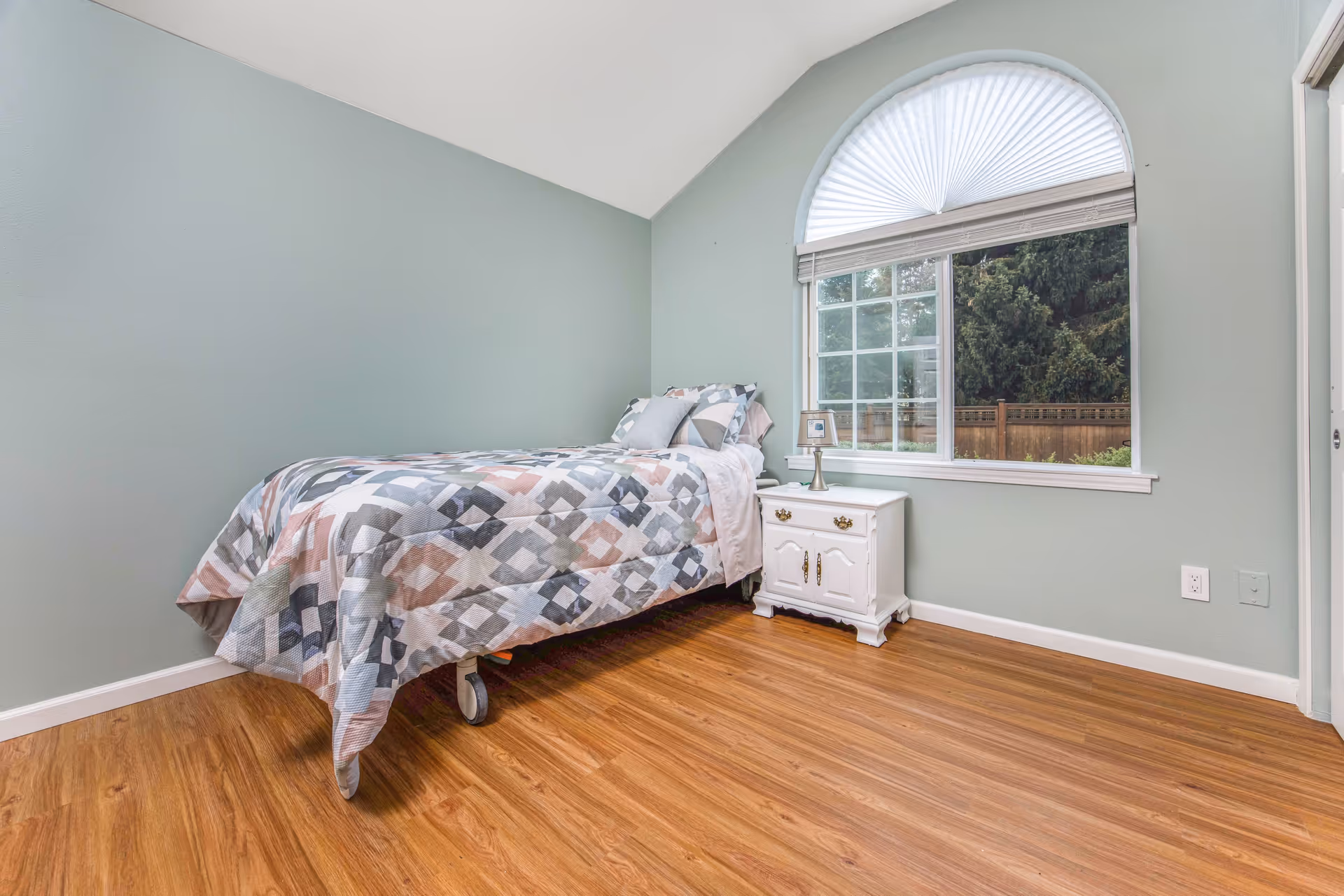 A simple bedroom with a single bed covered in a geometric patterned quilt in shades of gray, pink, and white. The bed is positioned against a light green wall. Next to the bed is a white nightstand with a small lamp. There is a large window with a white blind and a decorative half-circle fan shade above it, showing a view of trees and a wooden fence outside. The floor is wooden with a natural finish.