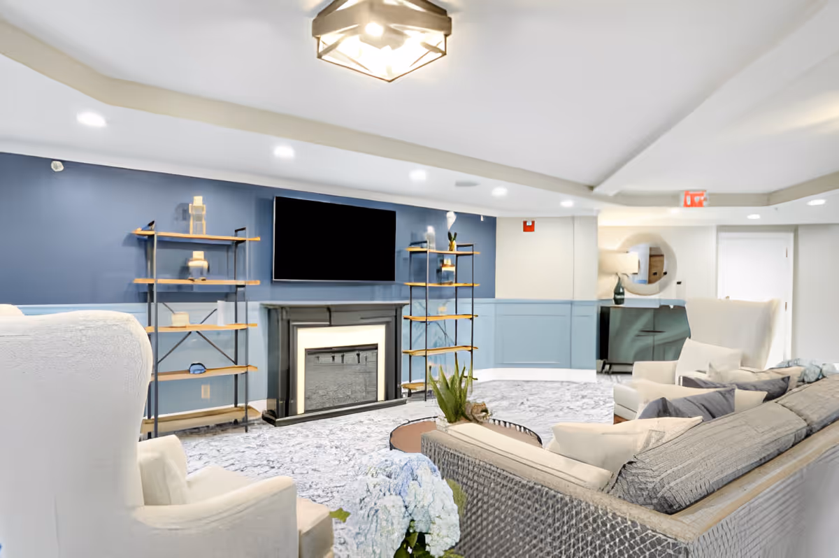 Bright communal living room with a wall-mounted TV over a fireplace, shelving, and upholstered seating.