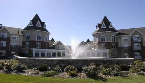 Exterior view of a large senior living facility with two prominent turret-style towers, a central water fountain, landscaped garden beds, and a clear blue sky.
