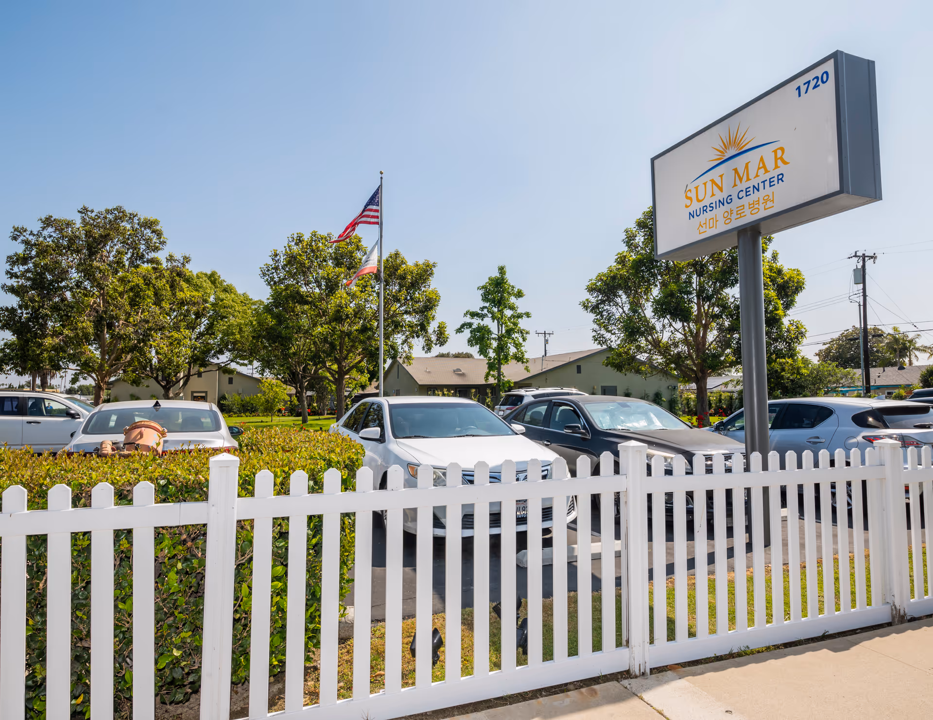 Exterior view of Sun Mar Nursing Center parking lot with several parked cars, a white picket fence in the foreground, trees, and two flagpoles with American and California flags under a clear blue sky.