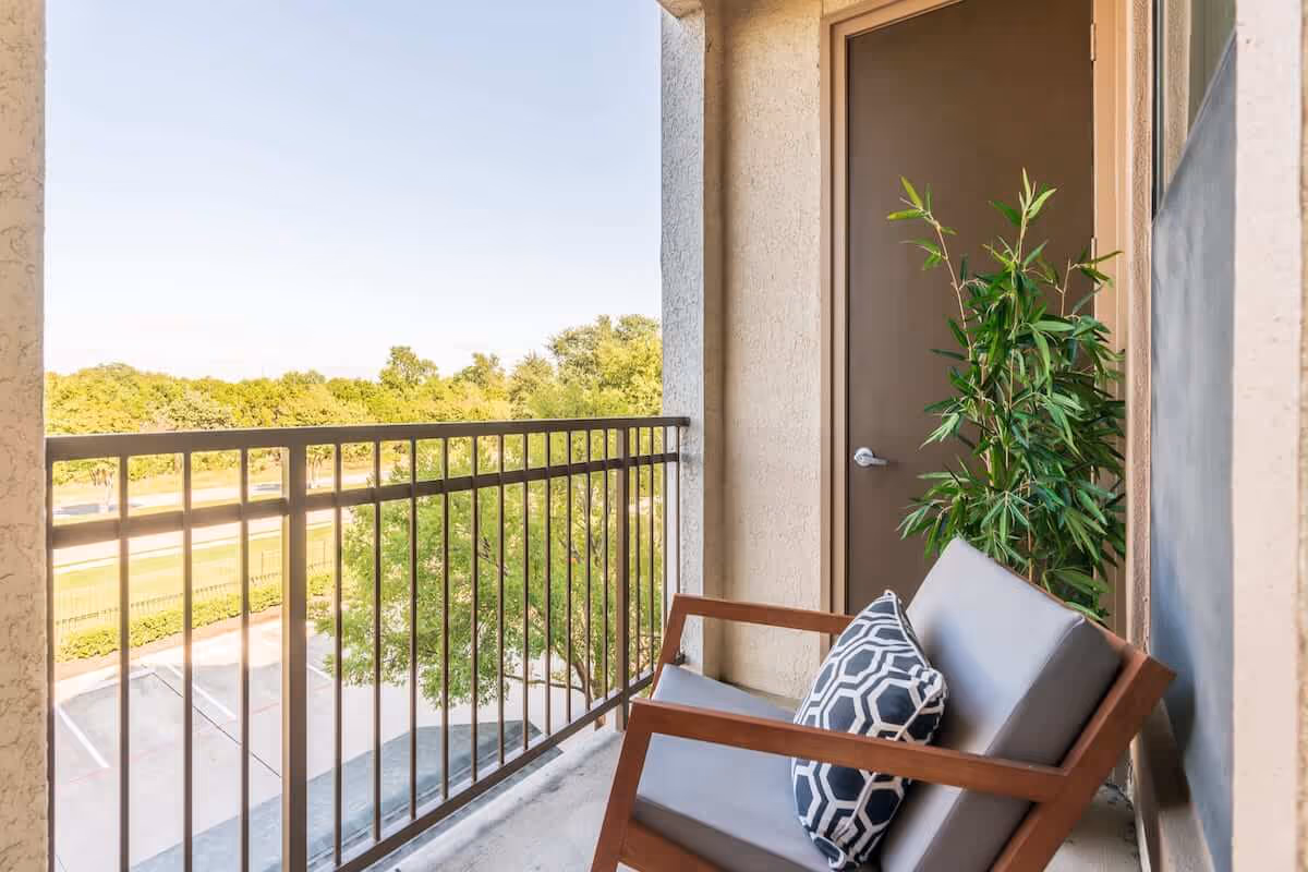 A small outdoor balcony with a wooden chair featuring gray cushions and a patterned pillow. There is a green potted plant next to a closed brown door. The balcony overlooks a parking area and green trees under a clear sky.