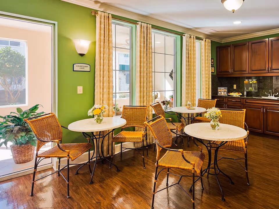 A cozy cafe area with green walls and large windows covered with yellow patterned curtains. There are three round tables with wicker chairs around them, each table decorated with a small vase of flowers. The floor is wooden, and there is a kitchen counter with dark wood cabinets and a sink in the background. A sign on the wall reads 'Courtyard Cafe'.