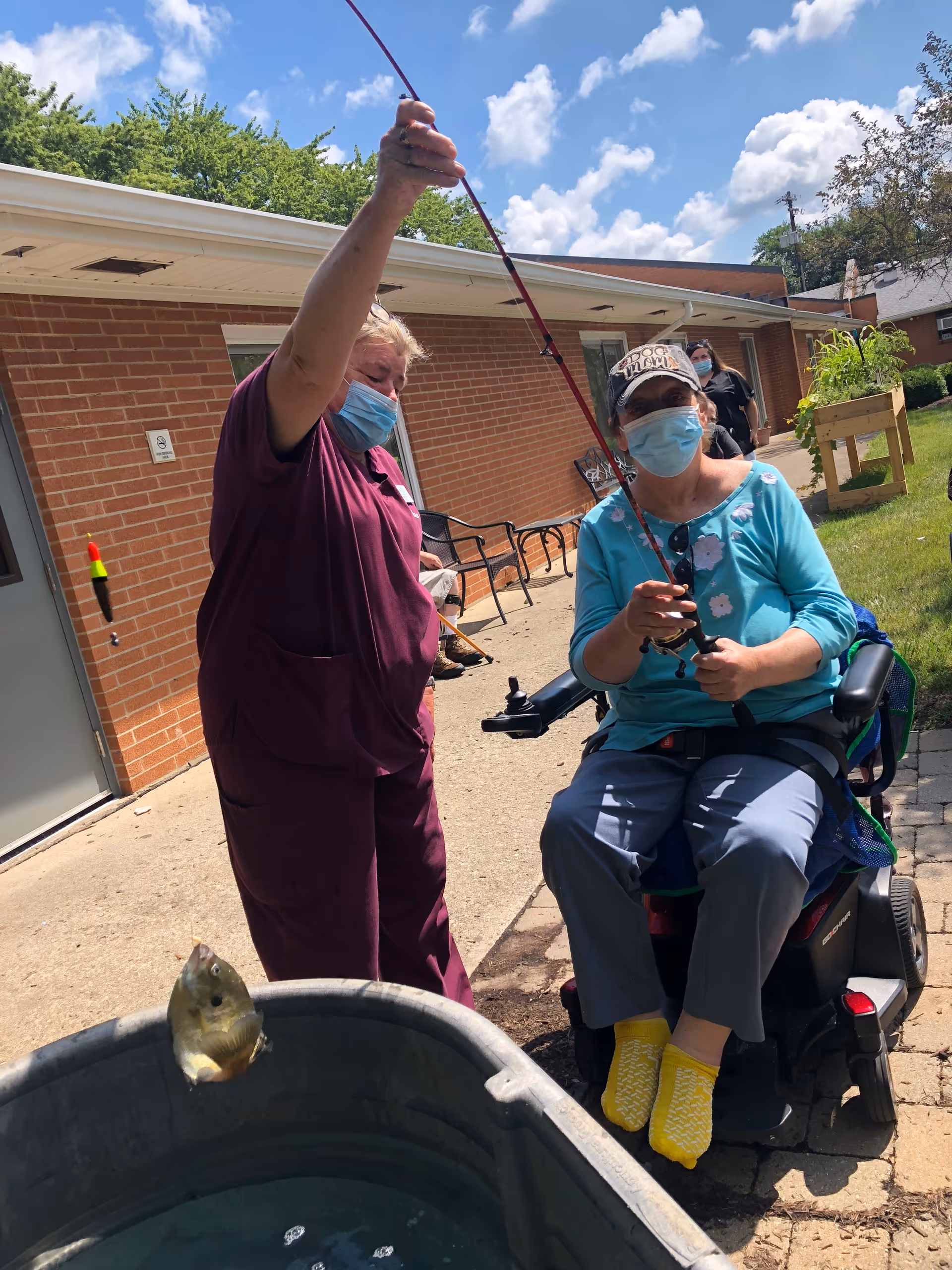 Two women wearing face masks are outdoors near a brick building. One woman in a maroon uniform is holding a fishing rod with a small fish caught on the line, while the other woman, seated in a motorized wheelchair and wearing a blue shirt and yellow socks, is holding the fishing rod handle. There is a large metal tub filled with water in front of them, and the sky is partly cloudy.