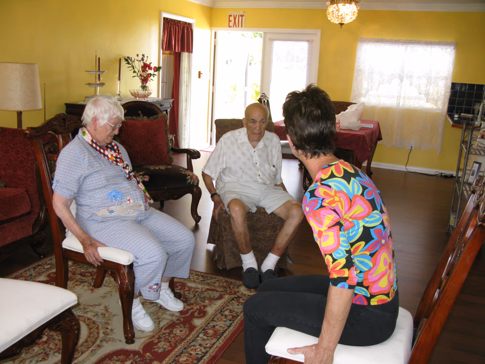 Three people sitting and conversing in a living room with yellow walls, wooden furniture, a patterned rug, and a window with lace curtains. The room has a warm and cozy atmosphere with an exit door visible in the background.