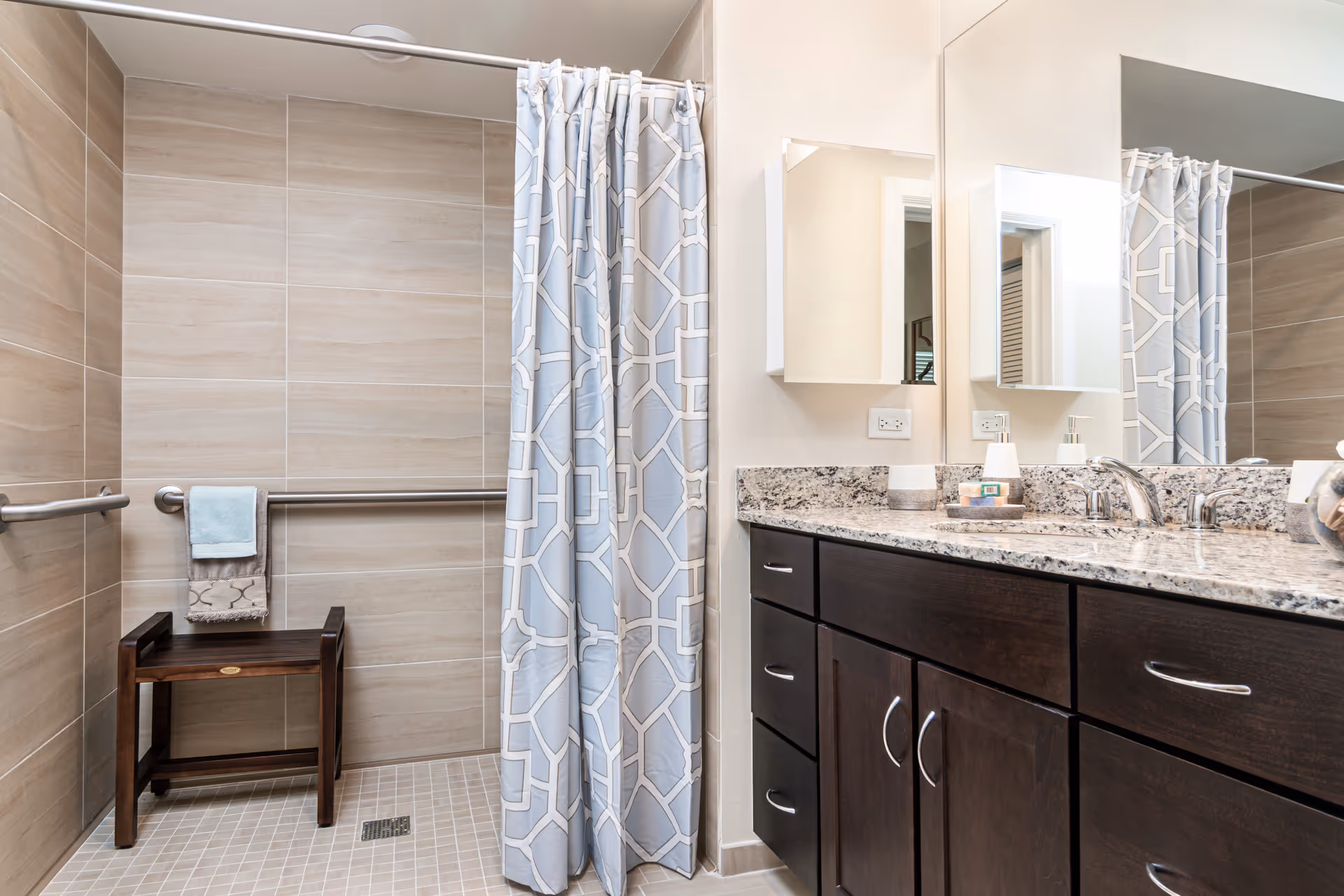 A modern bathroom featuring a walk-in shower with beige tiles, a silver grab bar, a wooden bench, and a blue and white patterned shower curtain. To the right is a granite countertop with a sink, dark wood cabinets, a wall-mounted mirror, and a white medicine cabinet.