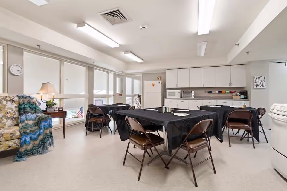 A bright room with multiple tables covered in black tablecloths surrounded by brown folding chairs. The room has a kitchenette area with white cabinets, a refrigerator, microwave, and toaster oven. A floral armchair with a colorful knitted blanket is positioned near a small side table with a lamp and a clock on the wall. Large windows let in natural light.