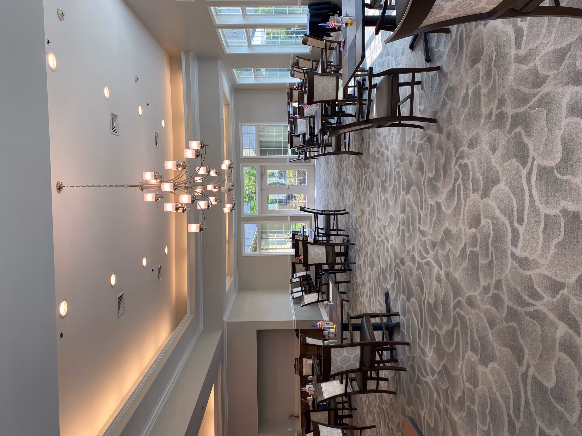 Spacious dining room with multiple tables and chairs, a chandelier, and large windows letting in natural light.