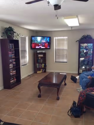 A cozy living room with tiled flooring, a wooden coffee table in the center, a flat-screen TV mounted on the wall between two windows with blinds, a tall bookshelf filled with DVDs and books on the left, and a glass display cabinet on the right. Two people are partially visible sitting on chairs on the right side of the image. There are plants on top of the bookshelf and display cabinet.