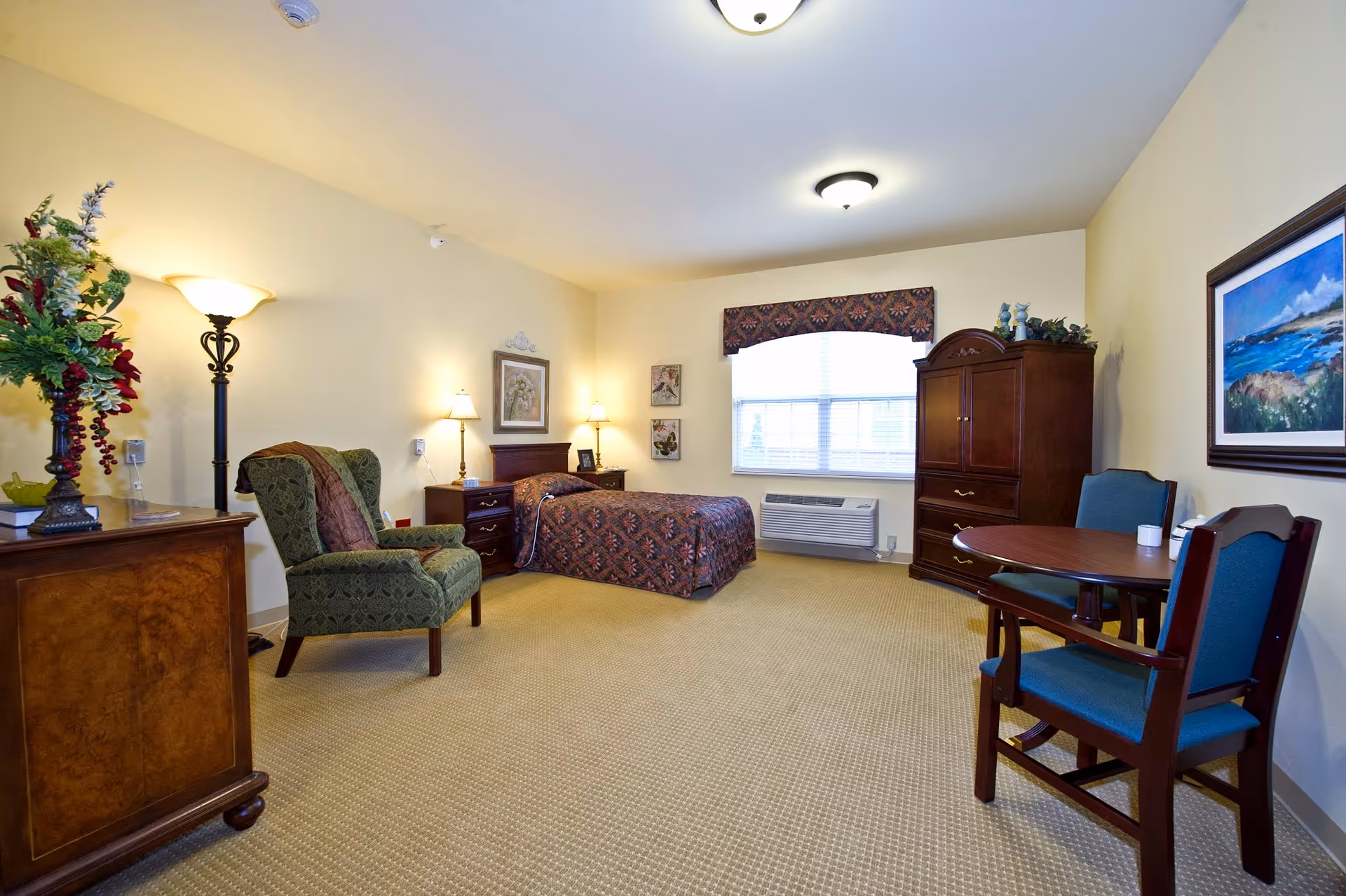 A well-lit bedroom in a senior living facility featuring a single bed with patterned bedding, two nightstands with lamps, a green upholstered armchair, a wooden dresser with a floral arrangement, a round wooden table with two blue cushioned chairs, a large window with a valance, and a wooden armoire. The walls are light-colored and decorated with framed artwork.