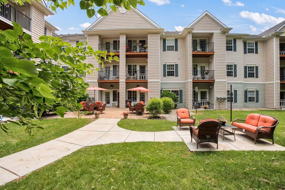 Outdoor patio area of a senior living facility with green grass, concrete walkways, and patio furniture including chairs and tables with umbrellas. The building has three stories with balconies and windows, surrounded by greenery and trees.