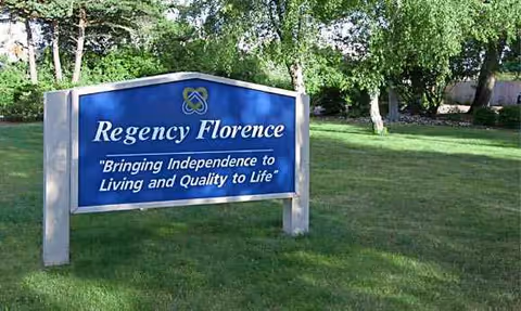 Blue Regency Florence sign on a grassy lawn with trees behind.