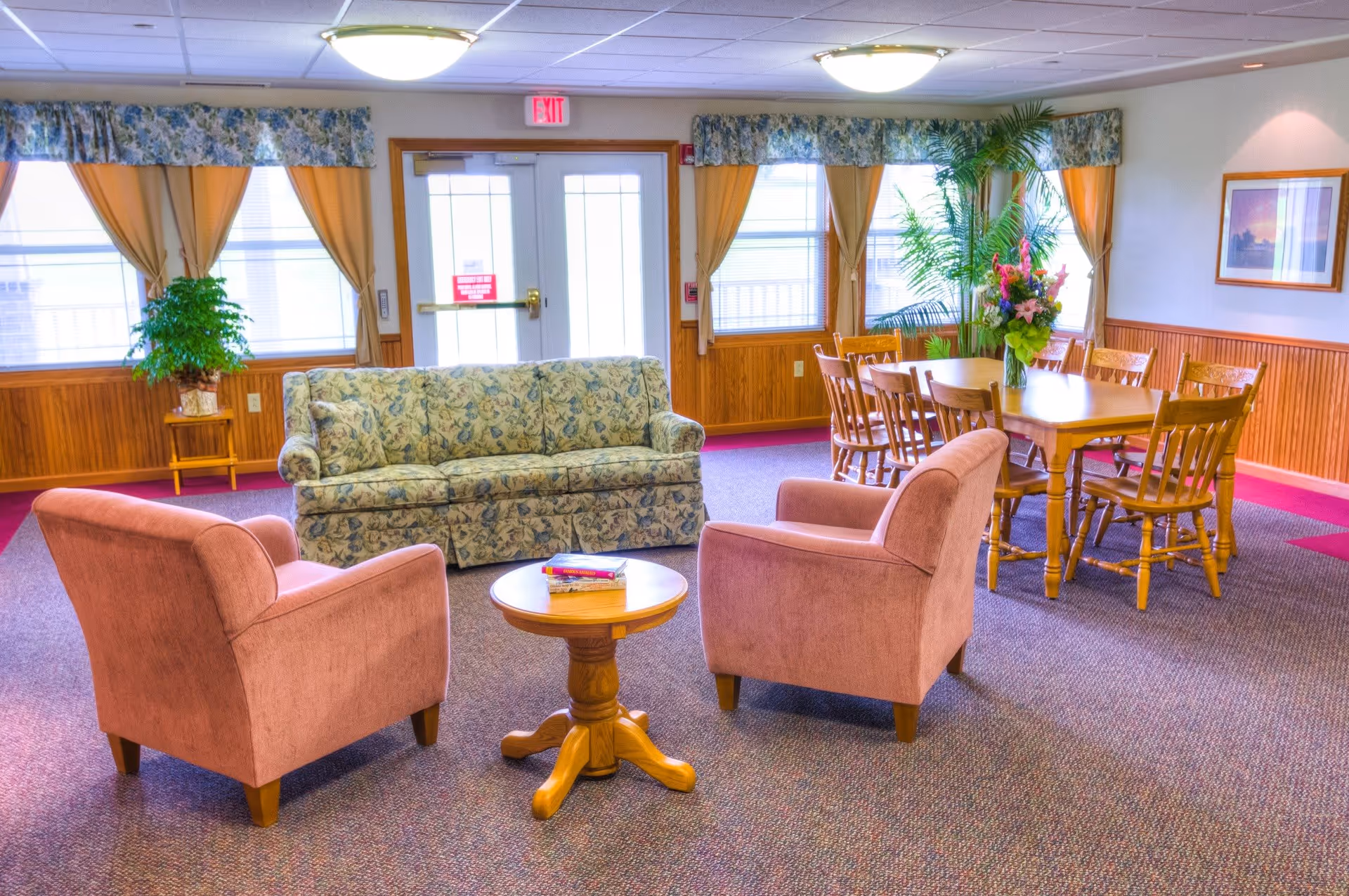 A bright and cozy common area with a floral-patterned sofa, two pink armchairs, and a small round wooden table with books on it. In the background, there is a wooden dining table with six chairs and a large floral arrangement. The room has large windows with yellow curtains and floral valances, wood paneling on the lower walls, and a framed picture on the wall.