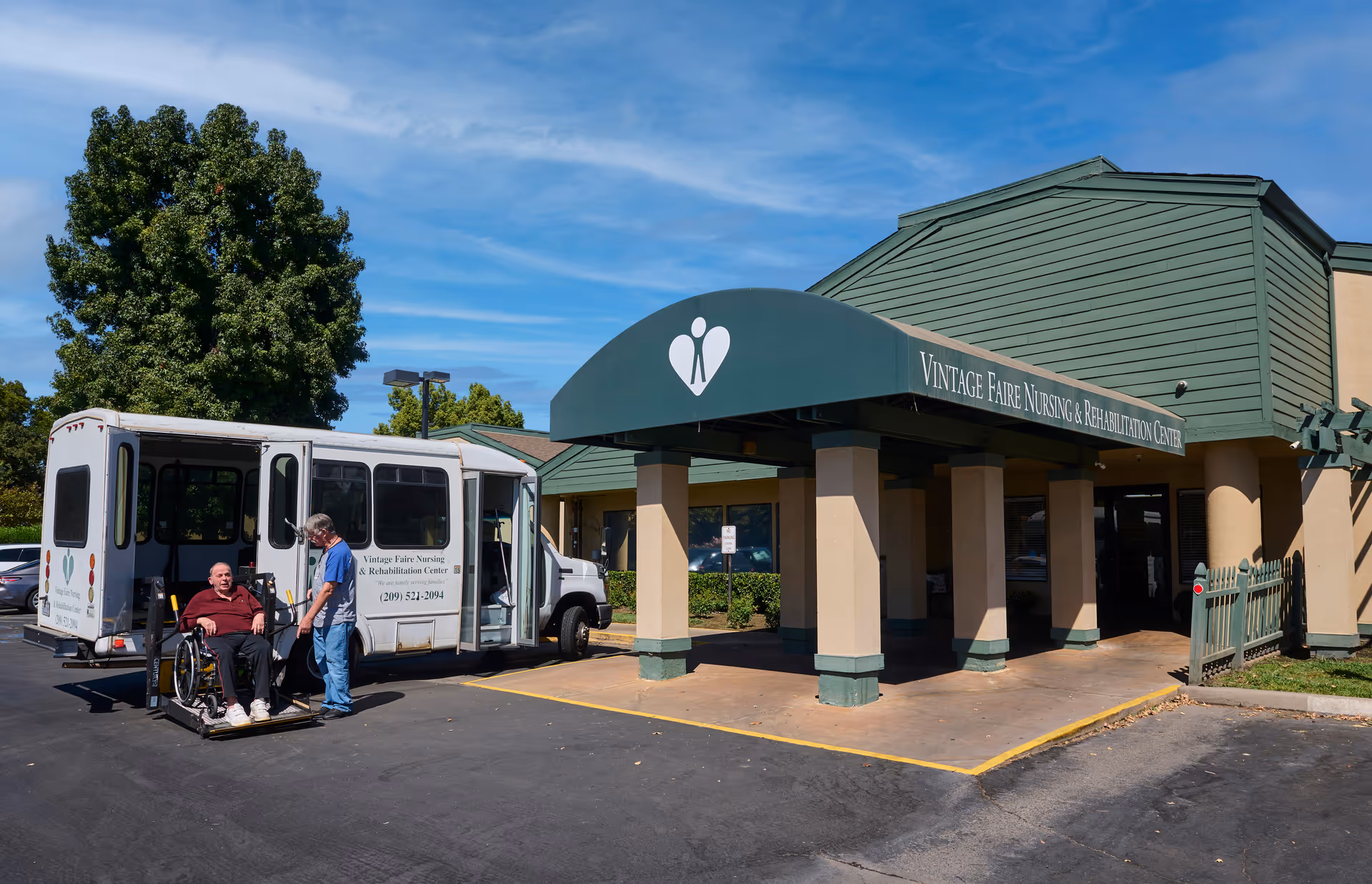 Front entrance of Vintage Faire Nursing & Rehabilitation Center with a shuttle van and a man in a wheelchair being assisted onto a lift.